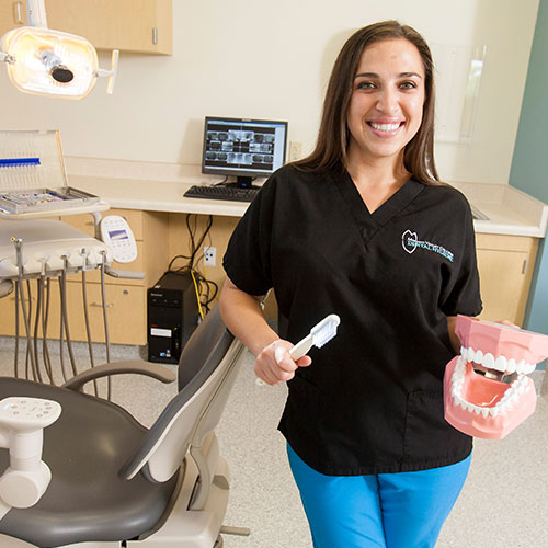 A dental hygiene student holds a model of teeth while standing next to dental hygiene equipment