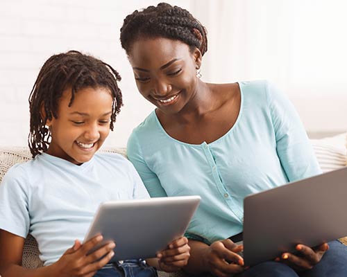 A mother does homework while her daughter uses a tablet