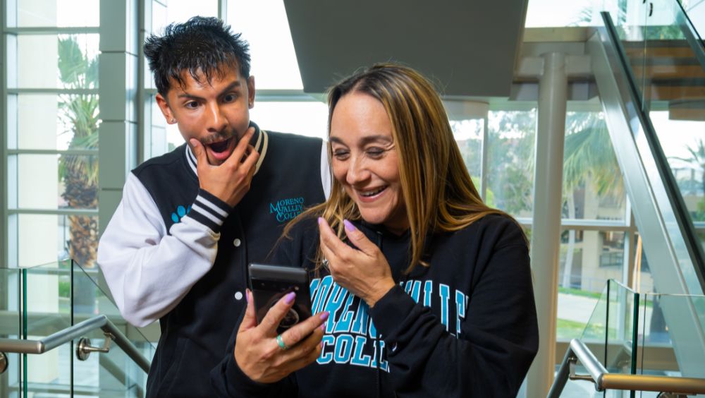 A Moreno Valley College staff member and student express delight and surprise while looking at a smartphone together in the Student Academic Services lobby.