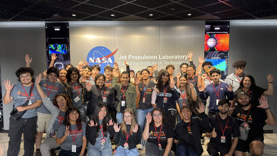 A large group of MESA students in program shirts and lanyards stand in front of the NASA JPL logo