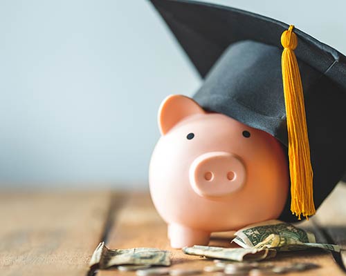 A pink piggy bank wears a graduation cap and sits next to a pile of change and dollar bills